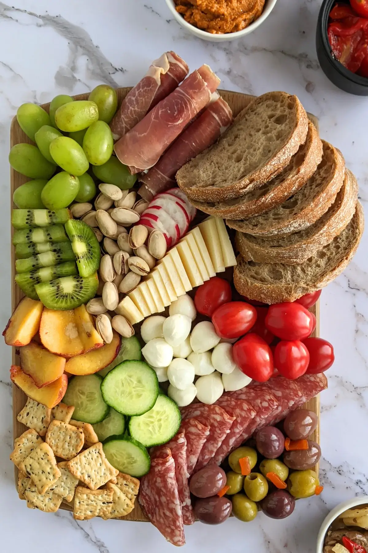 A vibrant colorful antipasto board with cured meats, marinated veggies, cheeses, olives, fruit, and ciabatta arranged on a wooden platter.