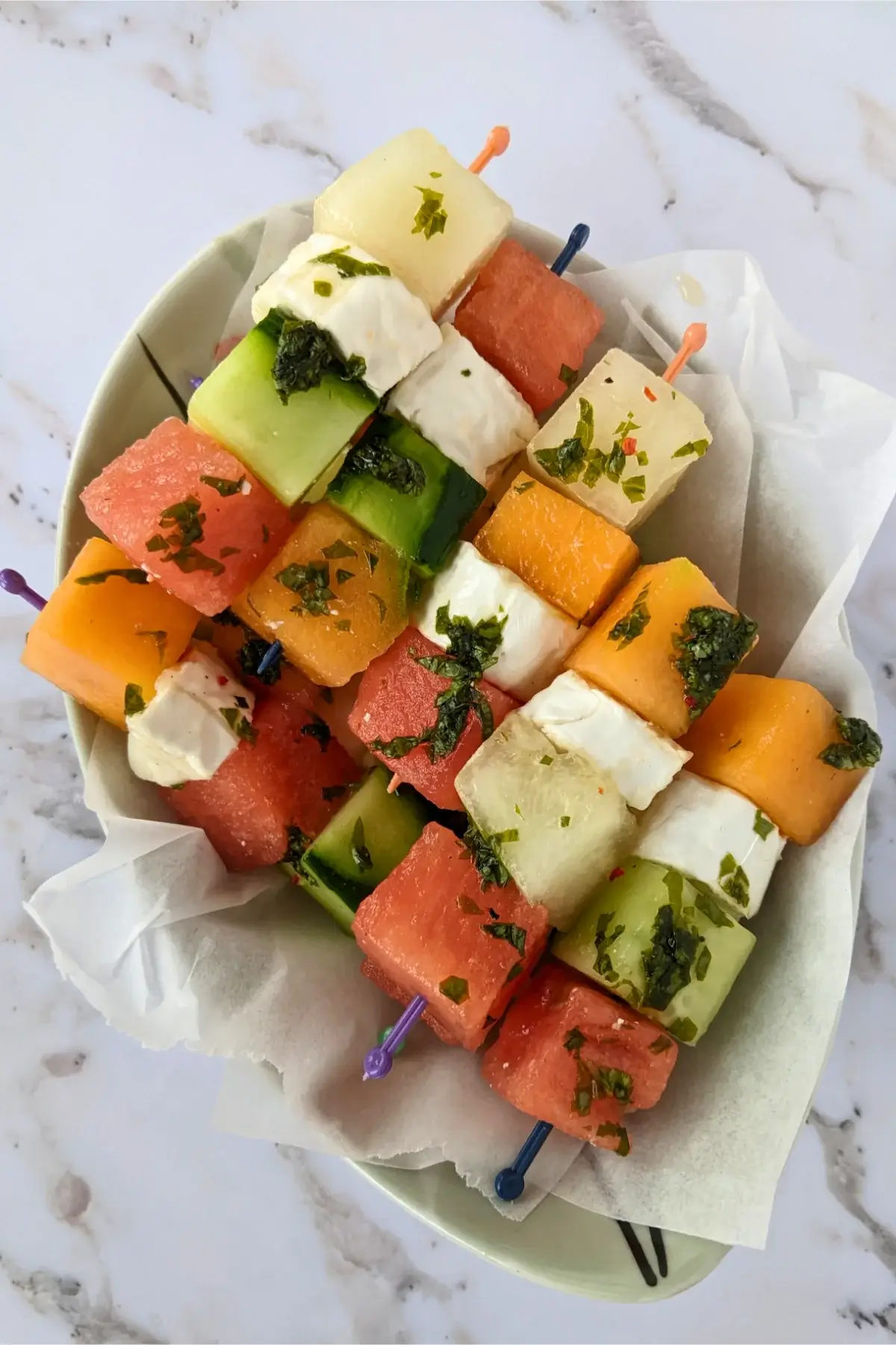 Colorful skewers with melon, watermelon, cucumber, and goat cheese drizzled with vinaigrette.