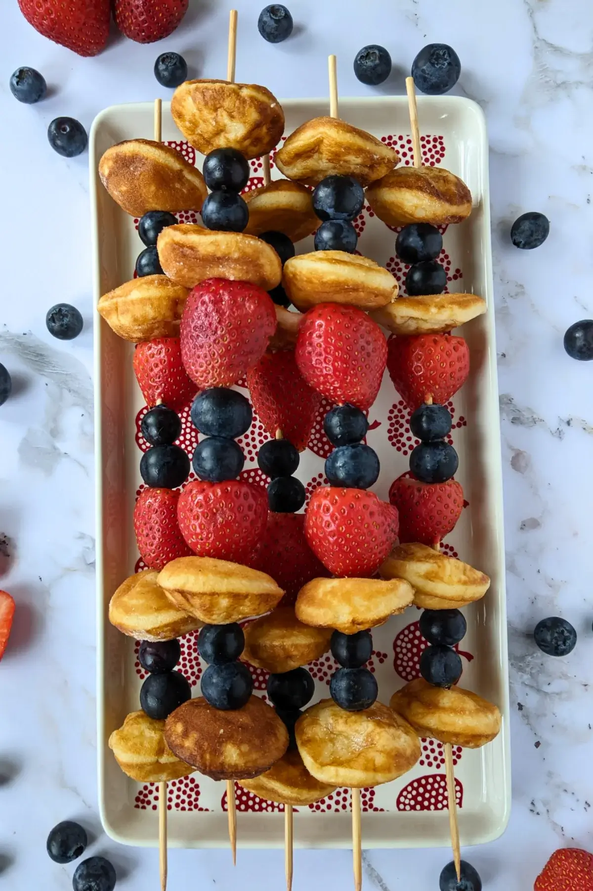 Mini pancake skewers alternating with blueberries and strawberries on wooden sticks.