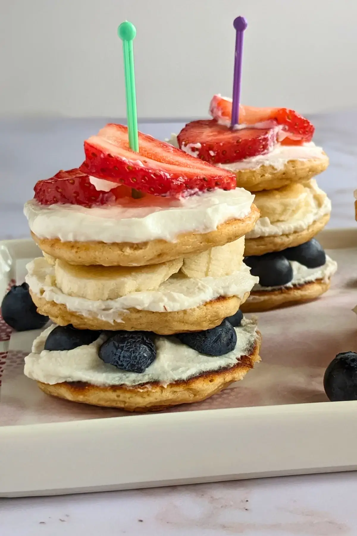 Patriotic mini pancake stacks layered with ricotta, banana slices, strawberries, and blueberries on a serving platter.