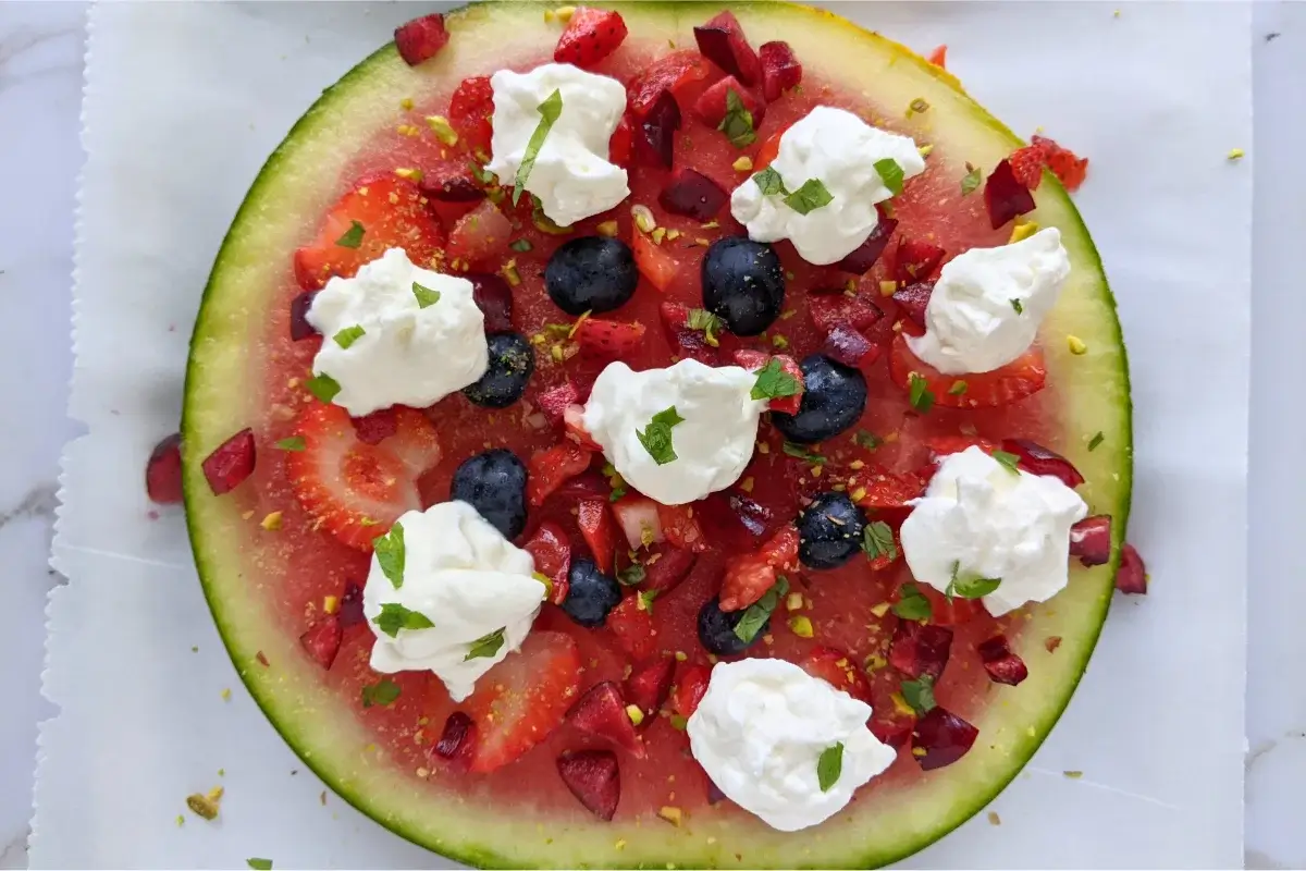 Watermelon slices topped like a pizza with whipped cream, strawberries, blueberries, cherries, and mint.