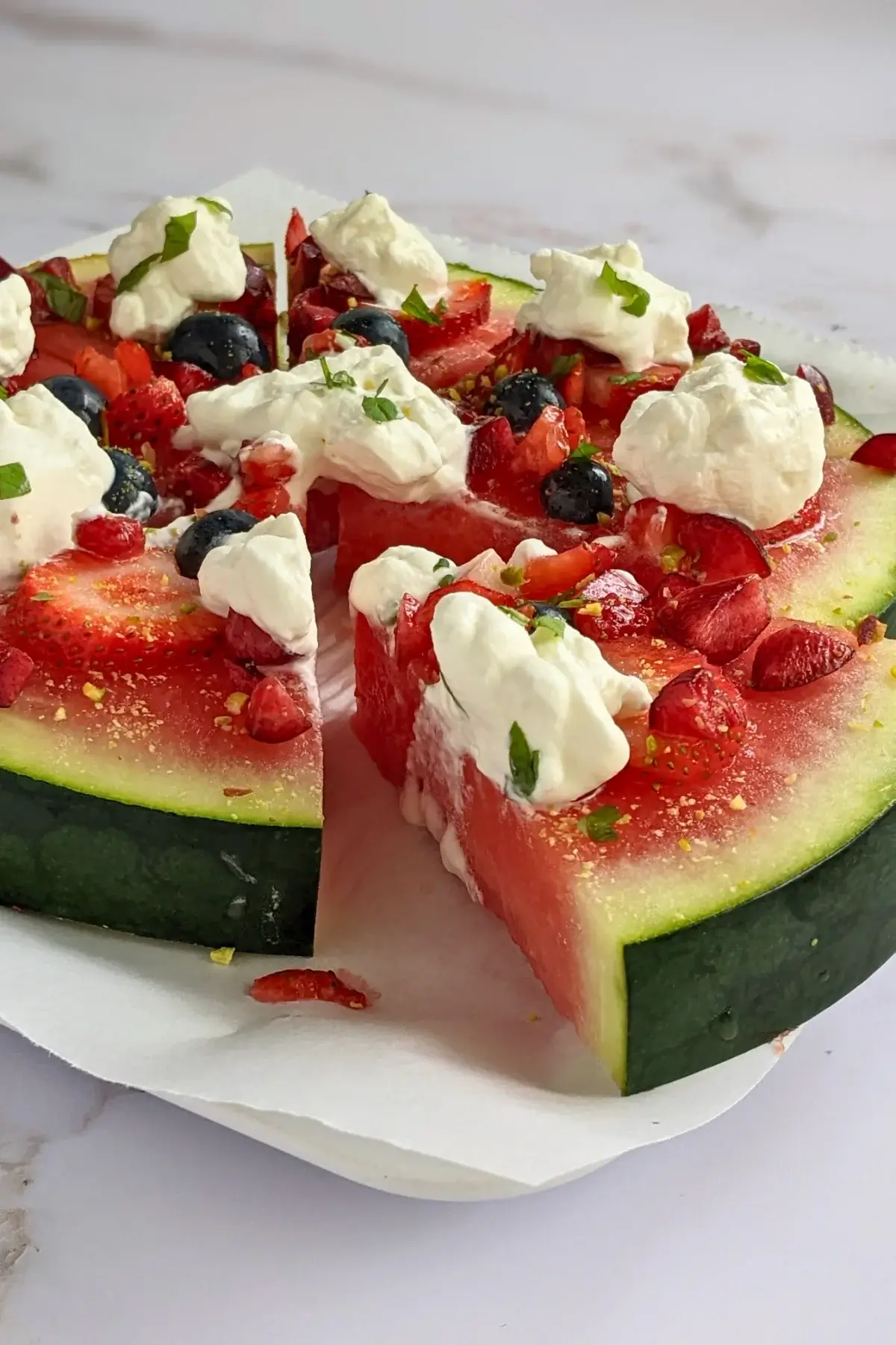 Watermelon pizza topped with blueberries, strawberries, cherries, whipped cream, and pistachios.