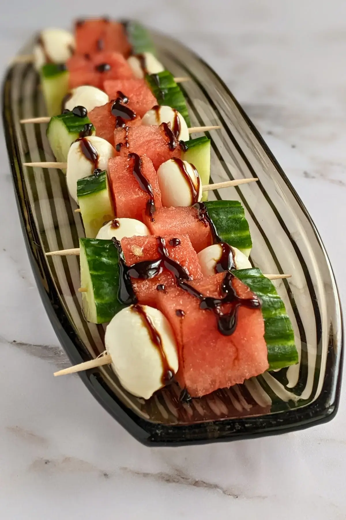 Watermelon, cucumber & mozzarella skewers drizzled with balsamic glaze on a white platter.