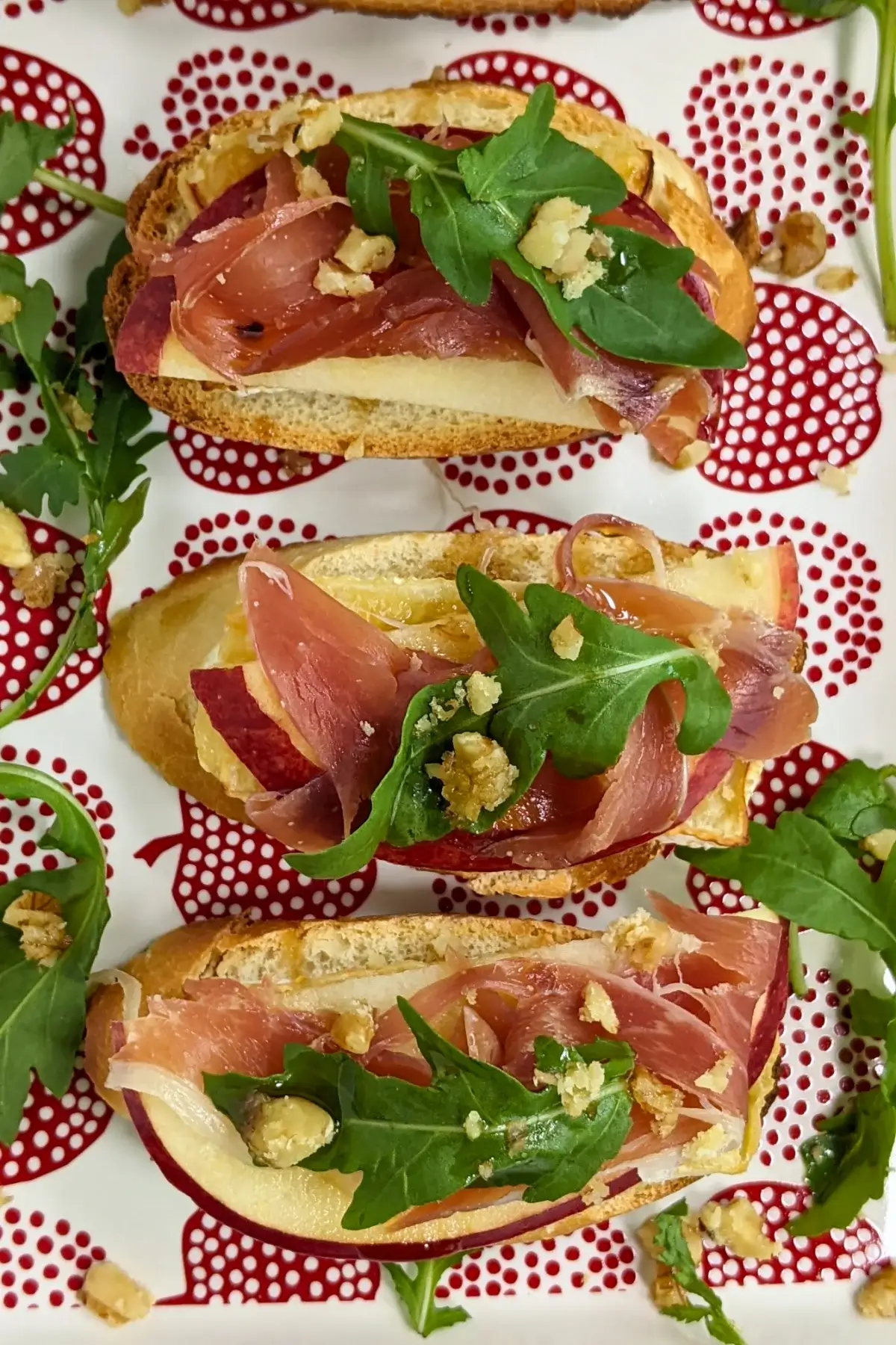 Crostini appetizers with apple, Brie cheese, and serrano ham topped with arugula, crushed walnuts and maple syrup.