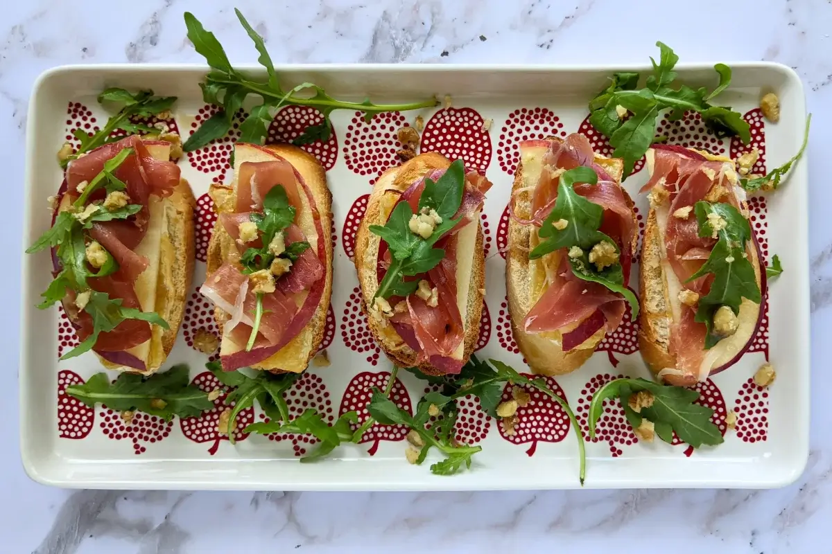 Crostini appetizers with apple, Brie cheese, and serrano ham topped with arugula, crushed walnuts and maple syrup.