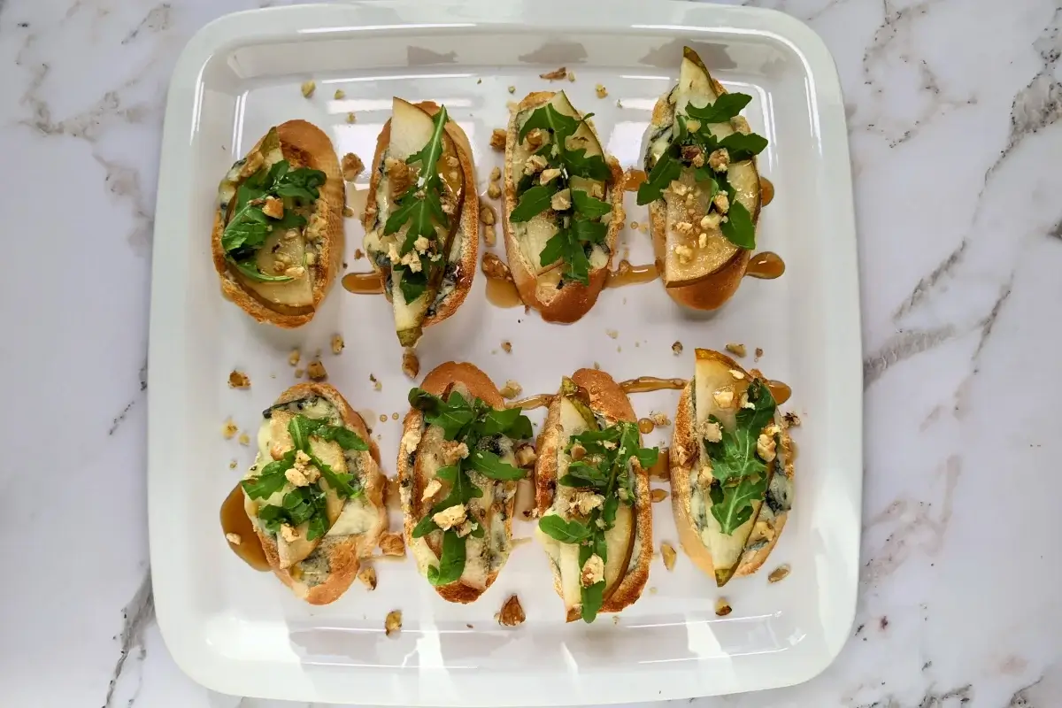 Crostini appetizers with pear and blue cheese, topped with arugula, crushed walnuts, and maple syrup.