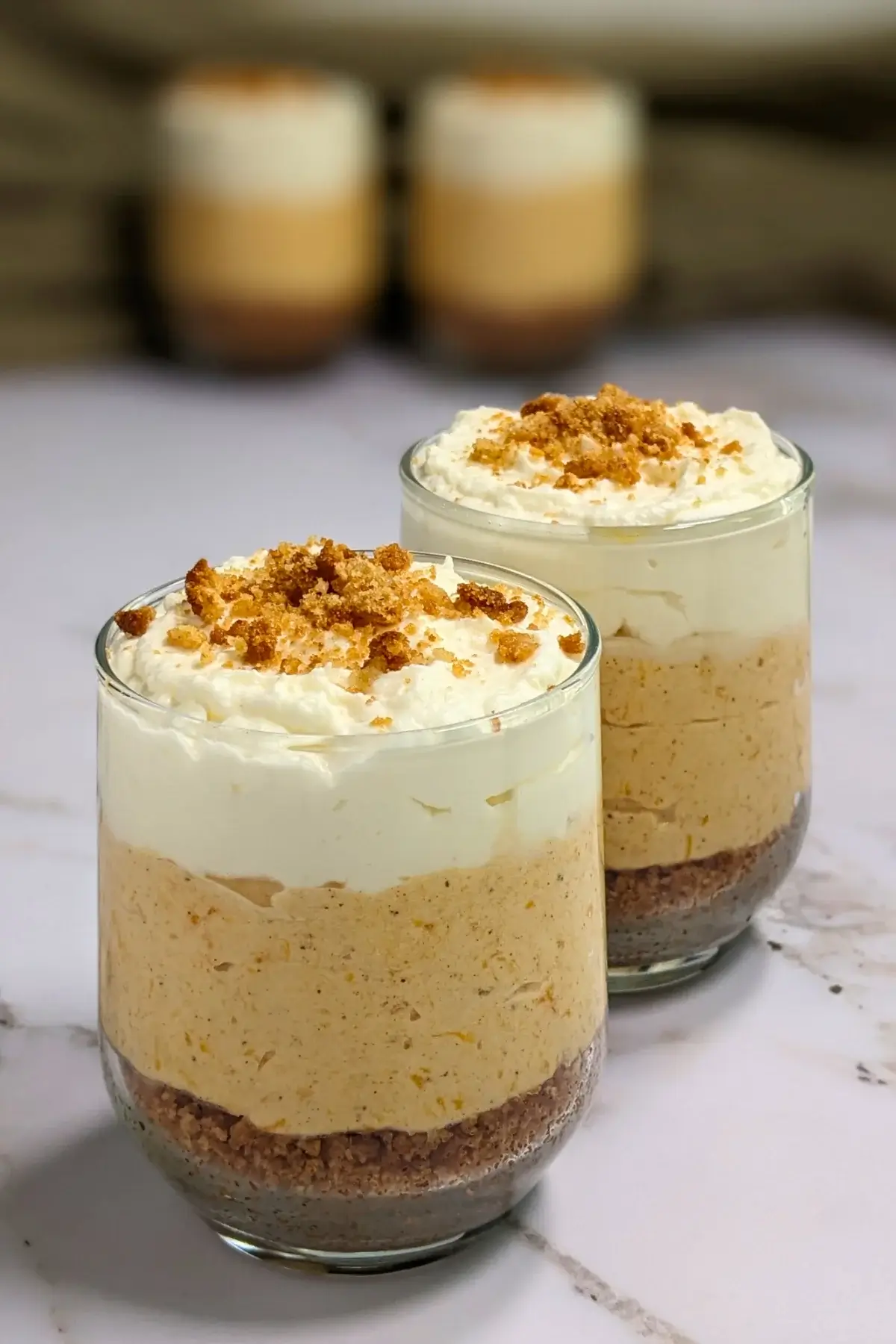 Pumpkin cheesecake mousse cups layered with a cookie and butter base; cream cheese, pumpkin, and whipped cream mousse; and topped with more whipped cream.