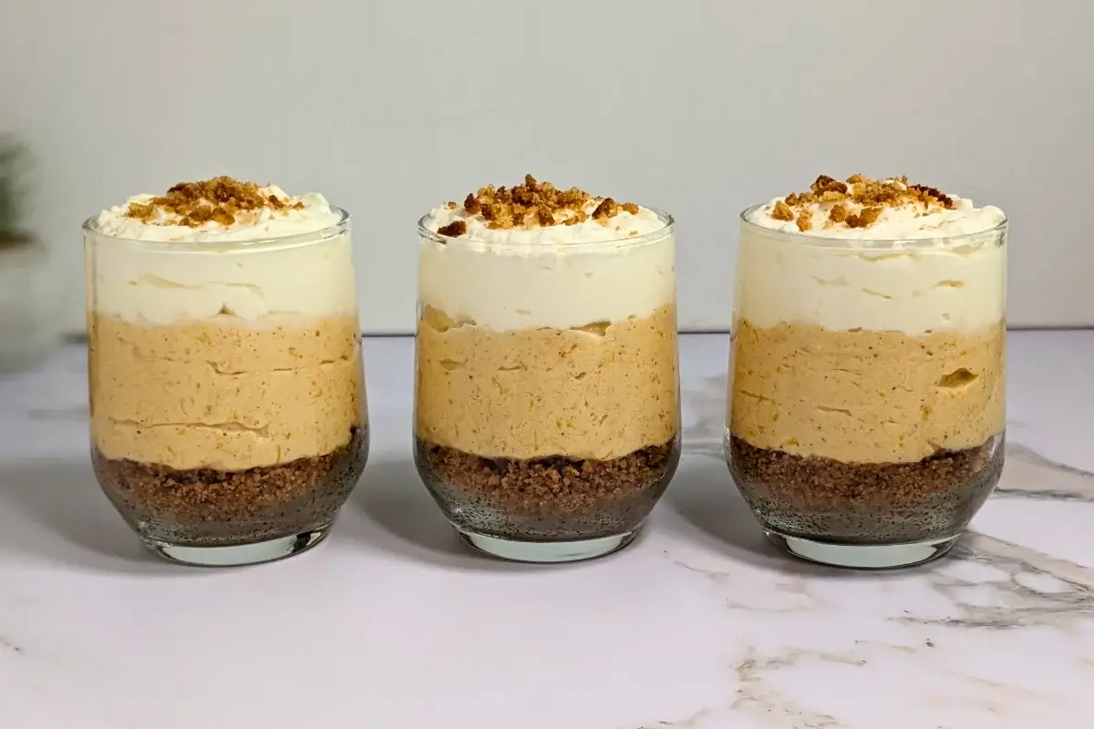 Pumpkin cheesecake mousse cups layered with a cookie and butter base; cream cheese, pumpkin, and whipped cream mousse; and topped with more whipped cream.