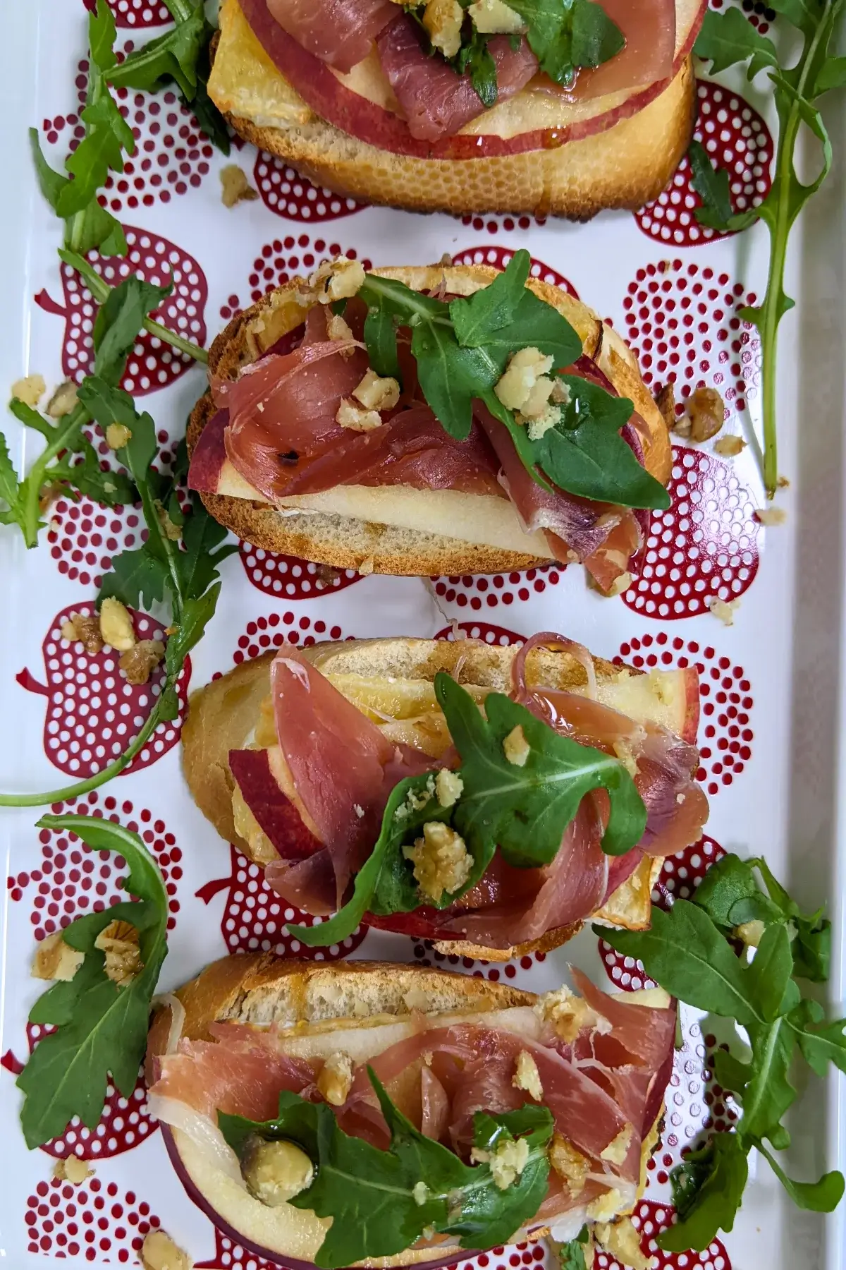 Crostini appetizers with apple, Brie cheese, and serrano ham topped with arugula, crushed walnuts and maple syrup.