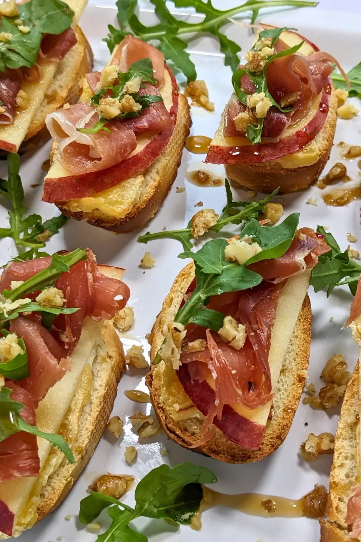 Crostini appetizers with apple, Brie cheese, and serrano ham topped with arugula, crushed walnuts and maple syrup.