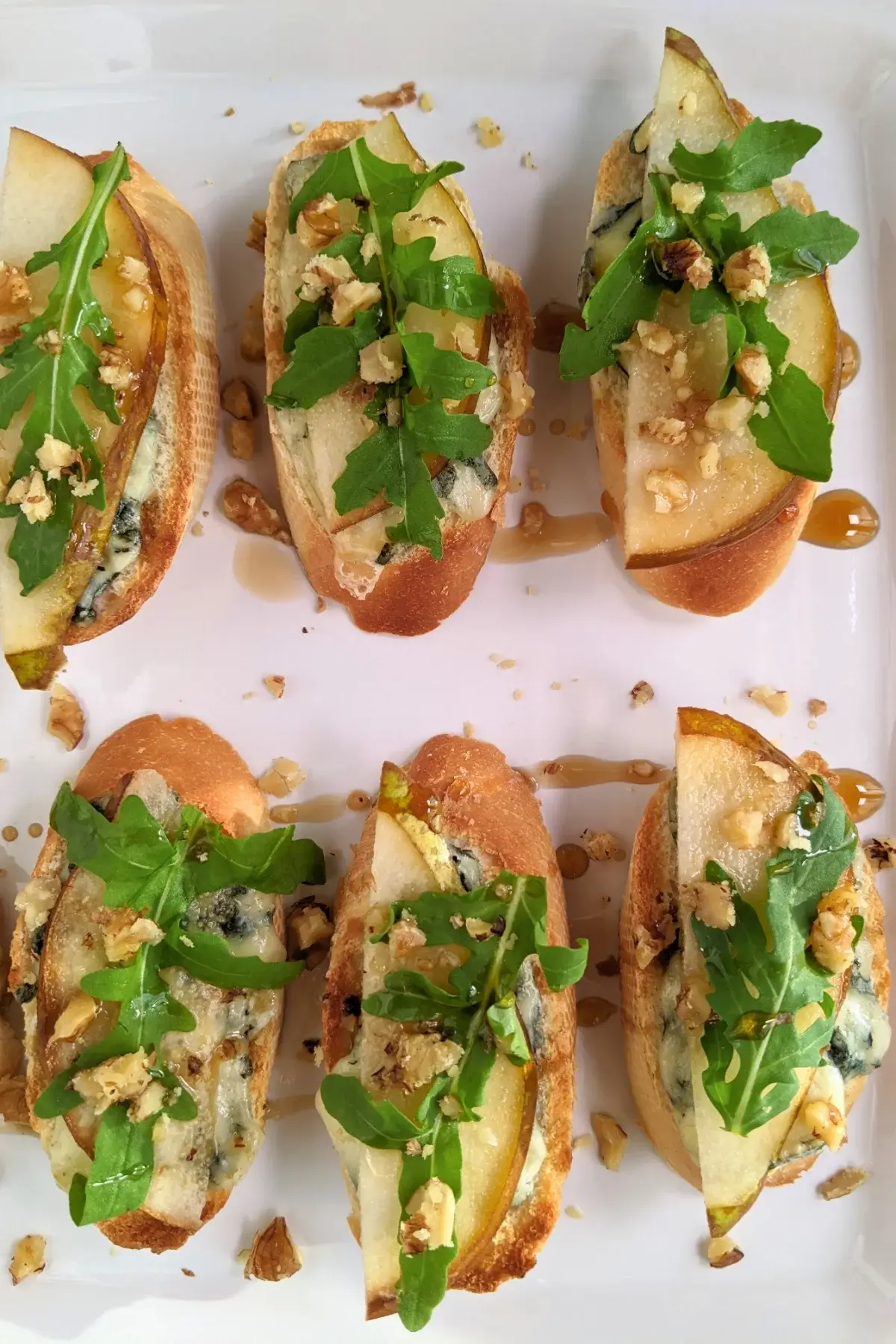 Crostini appetizers with pear and blue cheese, topped with arugula, crushed walnuts, and maple syrup.
