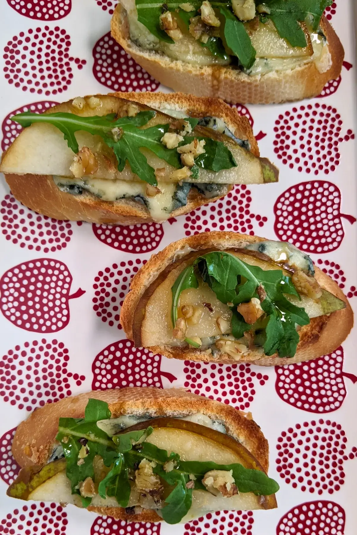 Crostini appetizers with pear and blue cheese, topped with arugula, crushed walnuts, and maple syrup.