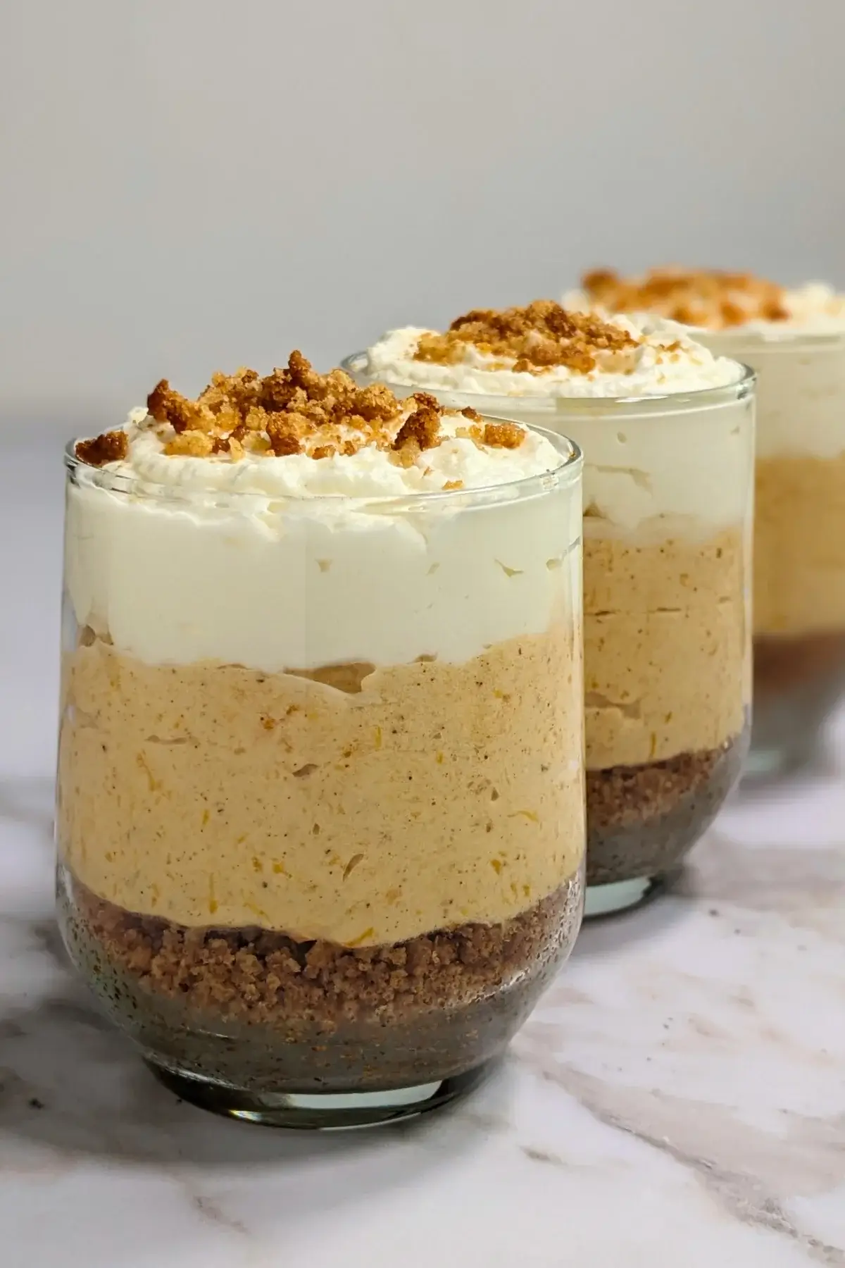 Pumpkin cheesecake mousse cups layered with a cookie and butter base; cream cheese, pumpkin, and whipped cream mousse; and topped with more whipped cream.